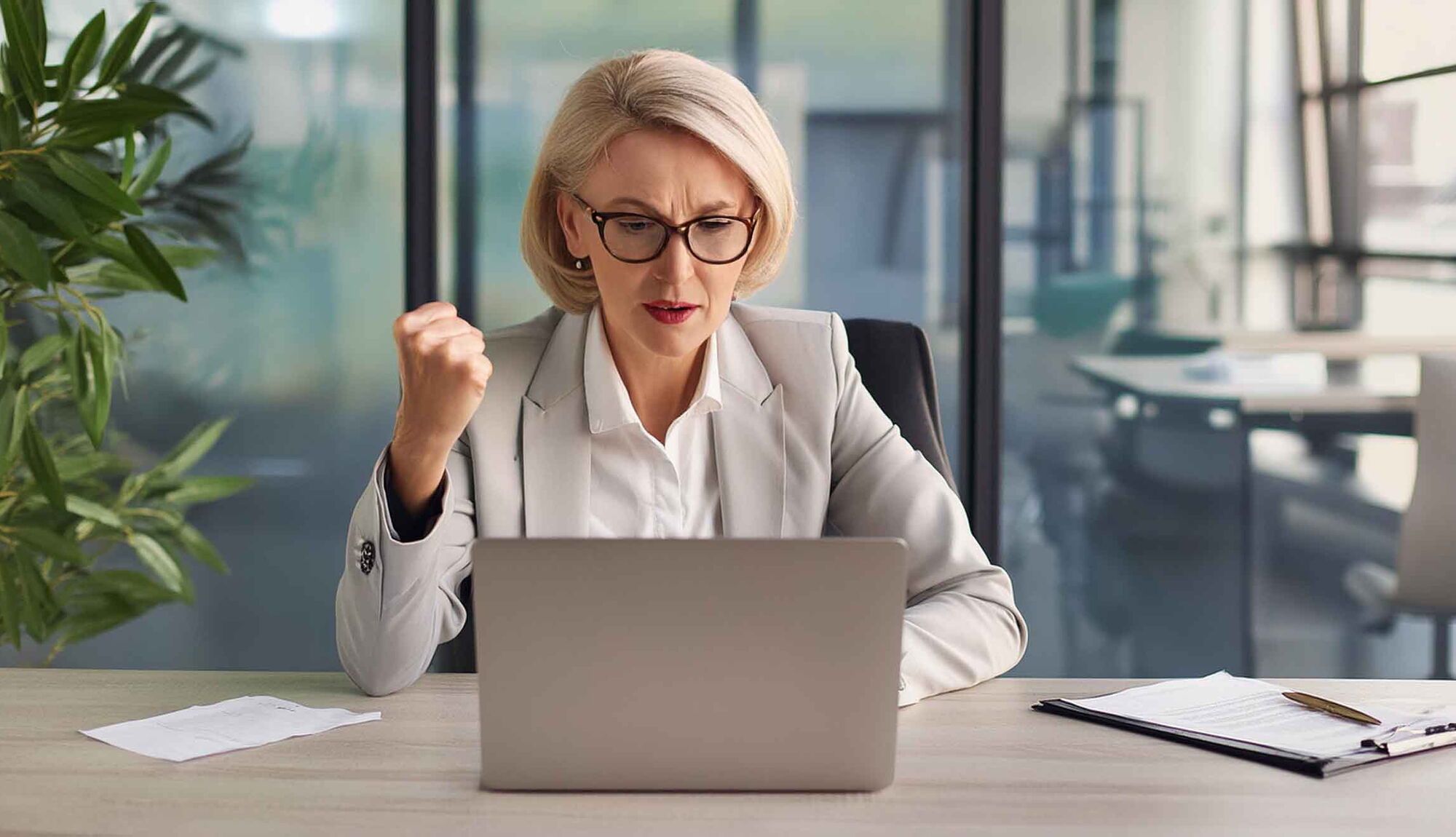 Businesswoman sits at her desk and waves her fist at her laptop.