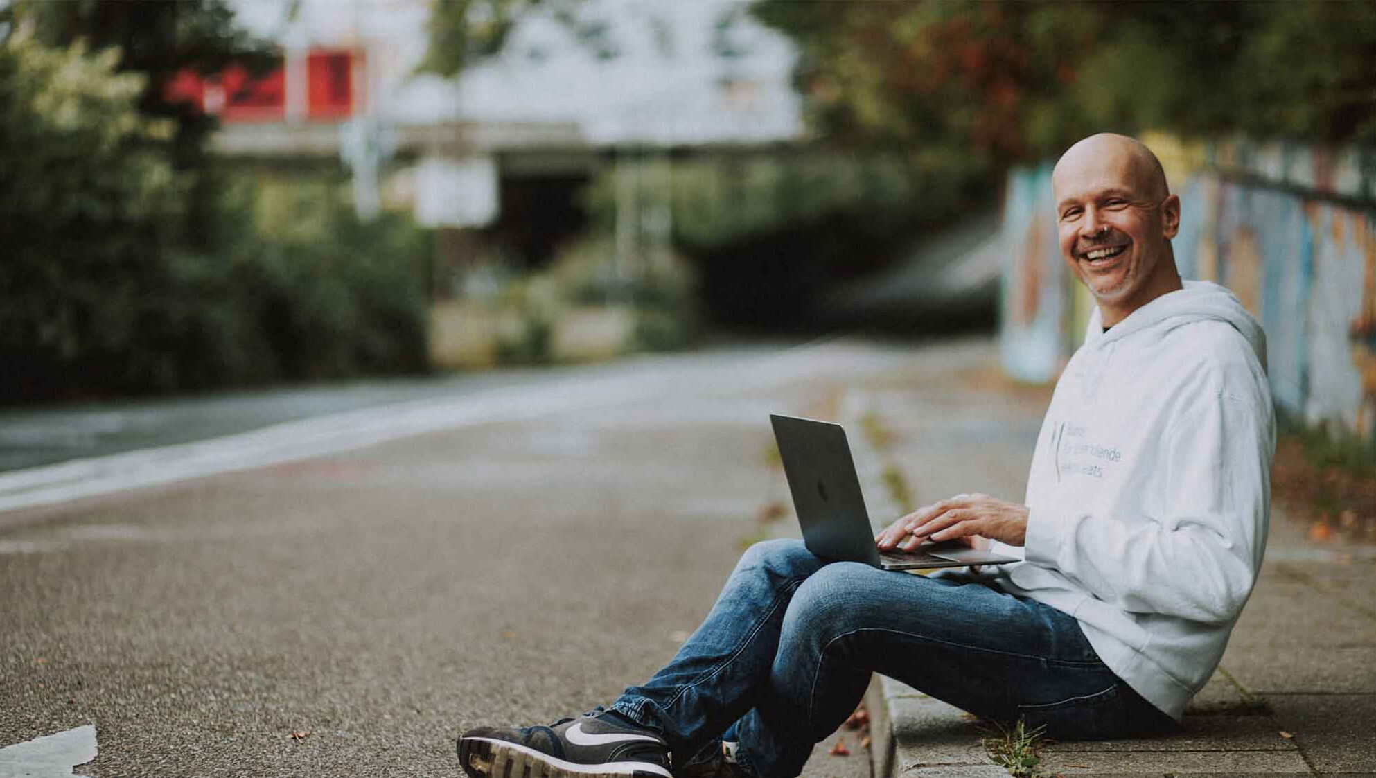 Webdesigner Paul Jackson sitzt am Bordstein mit Laptop und lächelt